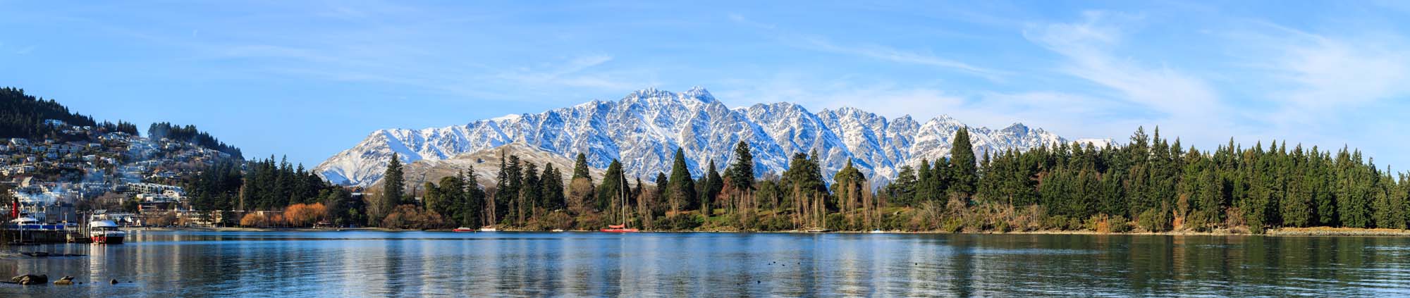 Queenstown Lake with Remarkables Mountains A serene lake view with snow-capped mountains in the background, featuring evergreen forests and a calm water surface in the foreground. The landscape showcases the dramatic natural beauty of New Zealand's South Island.