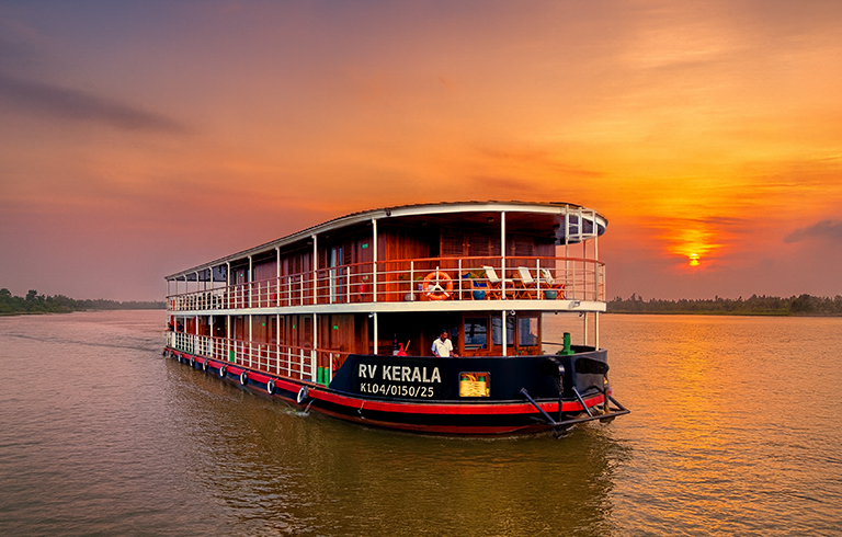 RV Kerala River Cruise at Sunset A multi-deck river cruise boat named RV Kerala sailing on a tranquil river during a vibrant orange sunset. The vessel reflects on the calm water with lush green landscape in the background.