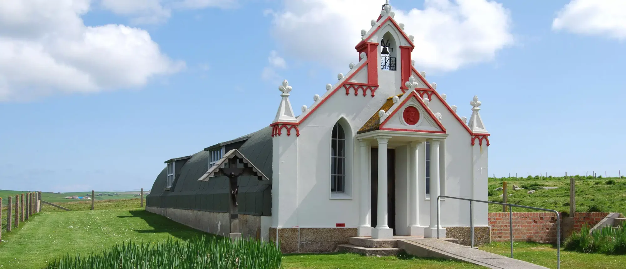 Red and White Chapel on Scottish Highland A charming small chapel featuring distinctive red and white Victorian architecture sits isolated in the rolling green hills of the Scottish Highlands, with a bell tower, pitched roof, and columned entrance framed by pastoral countryside.