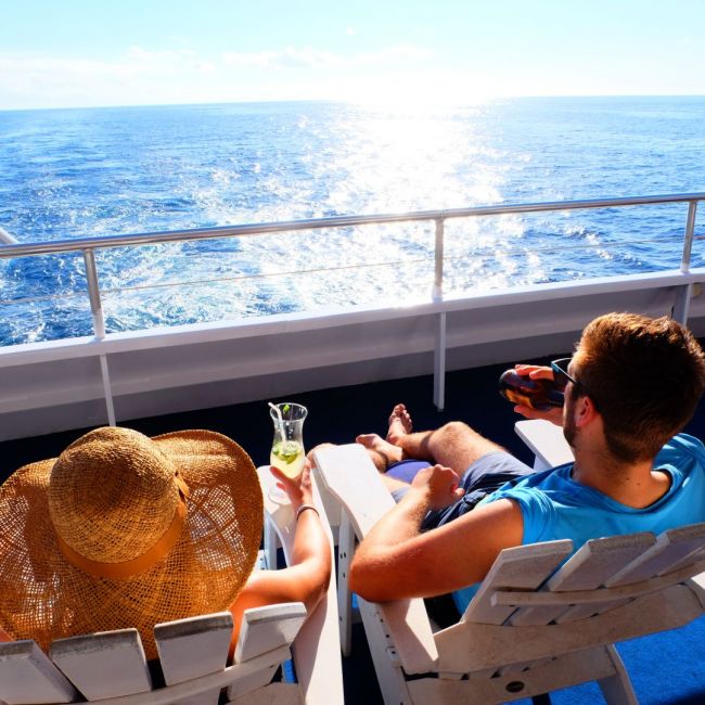 Relaxing on Cruise Ship Deck at Sea A passenger enjoys a leisurely afternoon on a cruise ship's deck, relaxing in a lounge chair with a refreshing beverage while gazing at the sparkling ocean waters. The bright sunlight reflects off the calm blue sea, creating a serene vacation atmosphere perfect for unwinding during a cruise holiday.