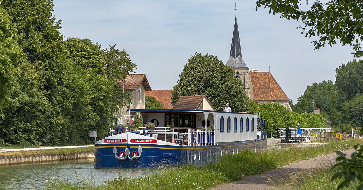 Renaissance River Cruise Boat in European Canal A Renaissance-branded river cruise boat with distinctive blue hull and white upper cabin navigates a serene European canal lined with lush green vegetation, with a charming village church featuring a pointed spire visible in the background.