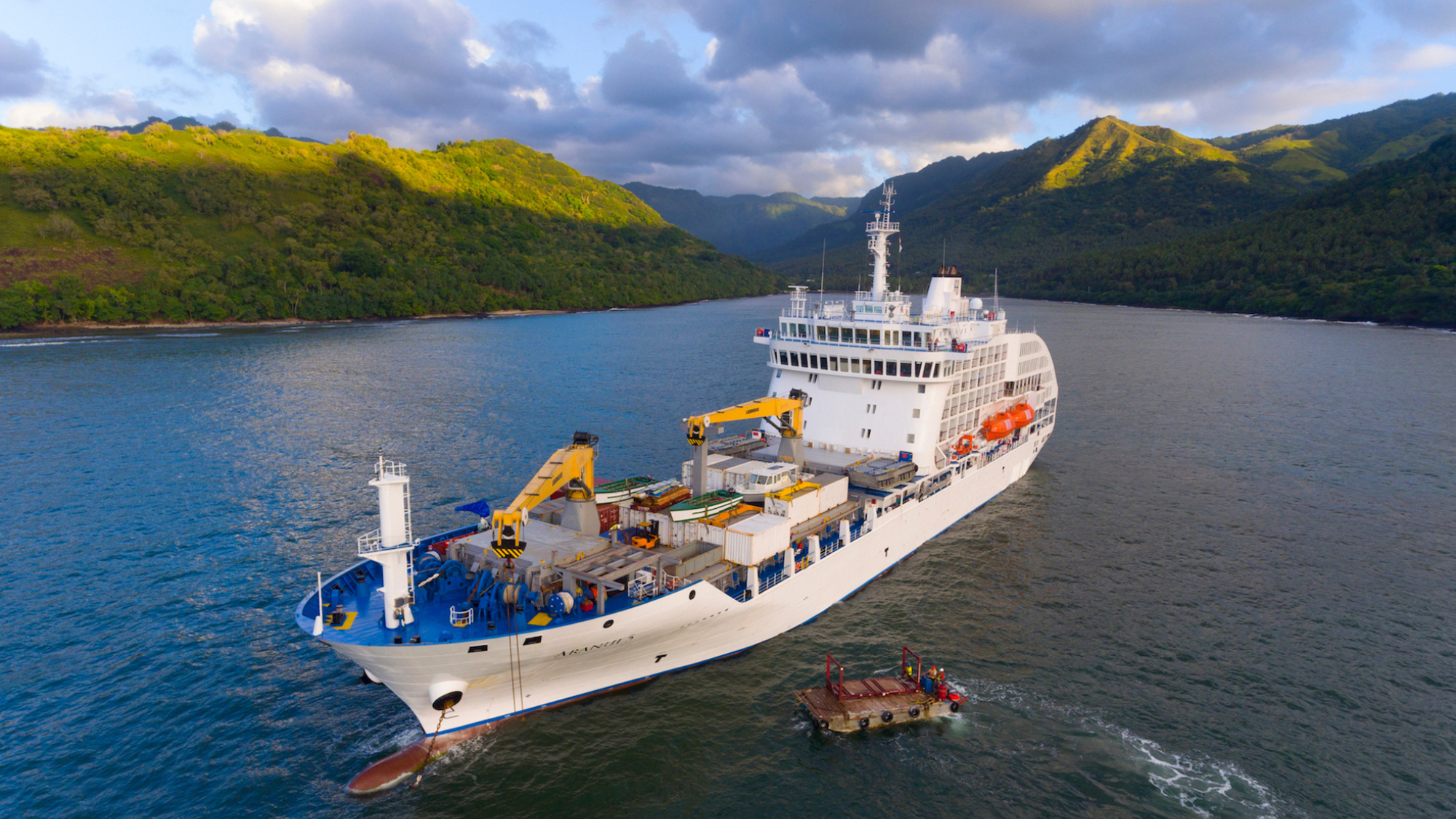 Research Vessel in Tropical Coastal Waters A white research or supply vessel navigating through a picturesque bay with lush green mountainous coastline, accompanied by a small support boat or platform.