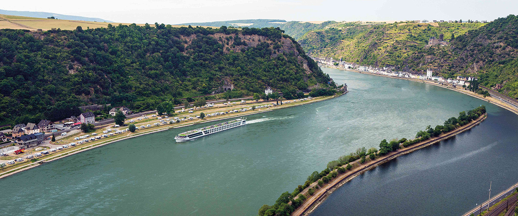 Rhine River Cruise Through Scenic Valley A sleek, modern white luxury yacht anchored near a historic coastal town with stone buildings perched on a green peninsula, surrounded by turquoise Mediterranean waters.