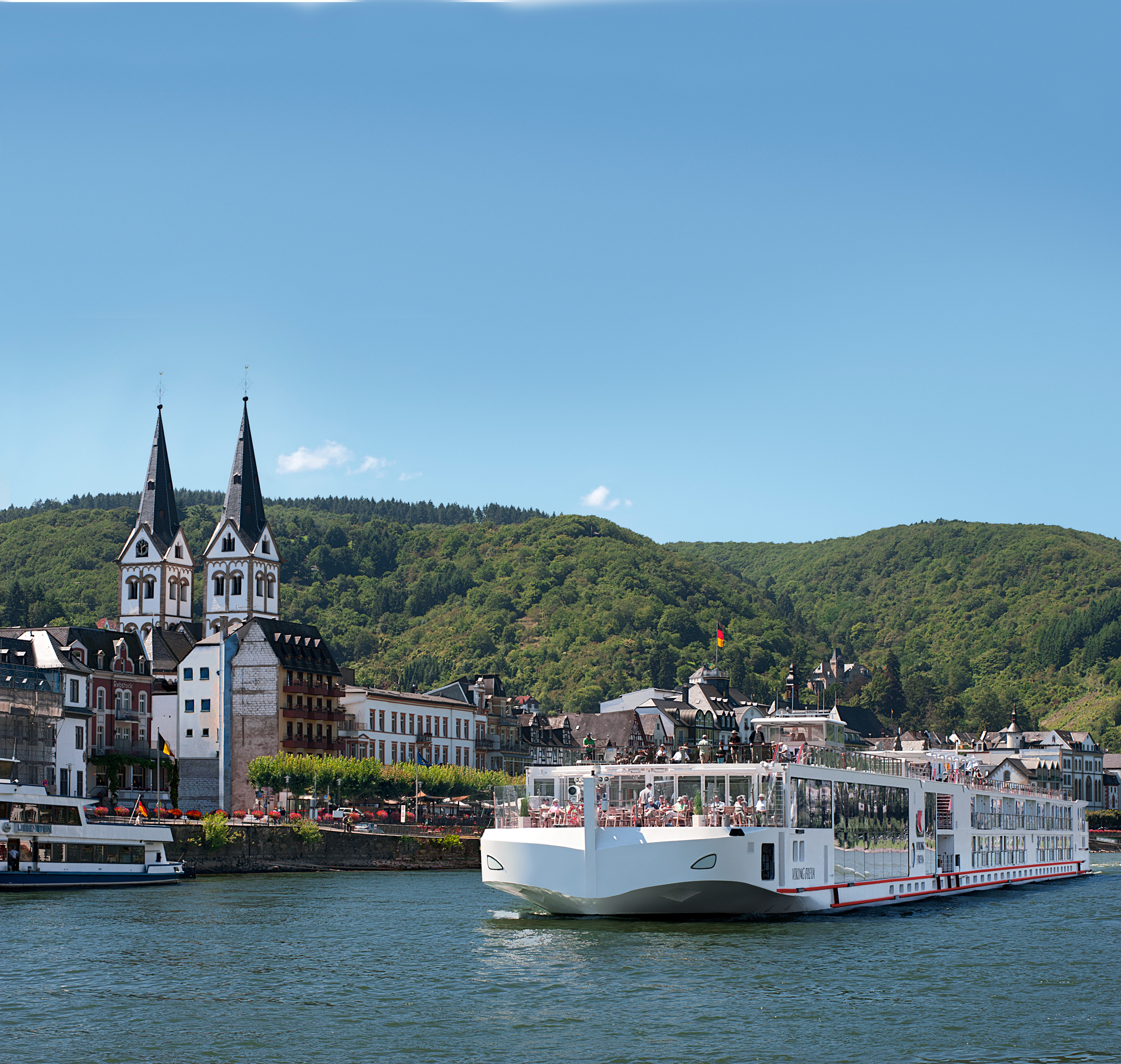 River Cruise Along the Moselle in Germany A white river cruise ship docked in a picturesque German town with twin-spired church towers, surrounded by lush green forested hills and a calm river.