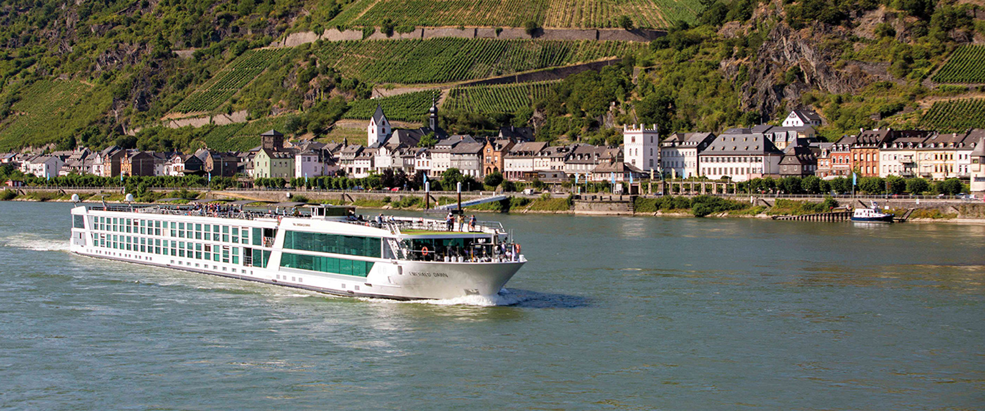 River Cruise Along the Rhine Valley A white and green river cruise ship sailing past a historic European town with vineyards and medieval buildings along the riverbank. The scene captures the scenic beauty of the Rhine River region in Germany.