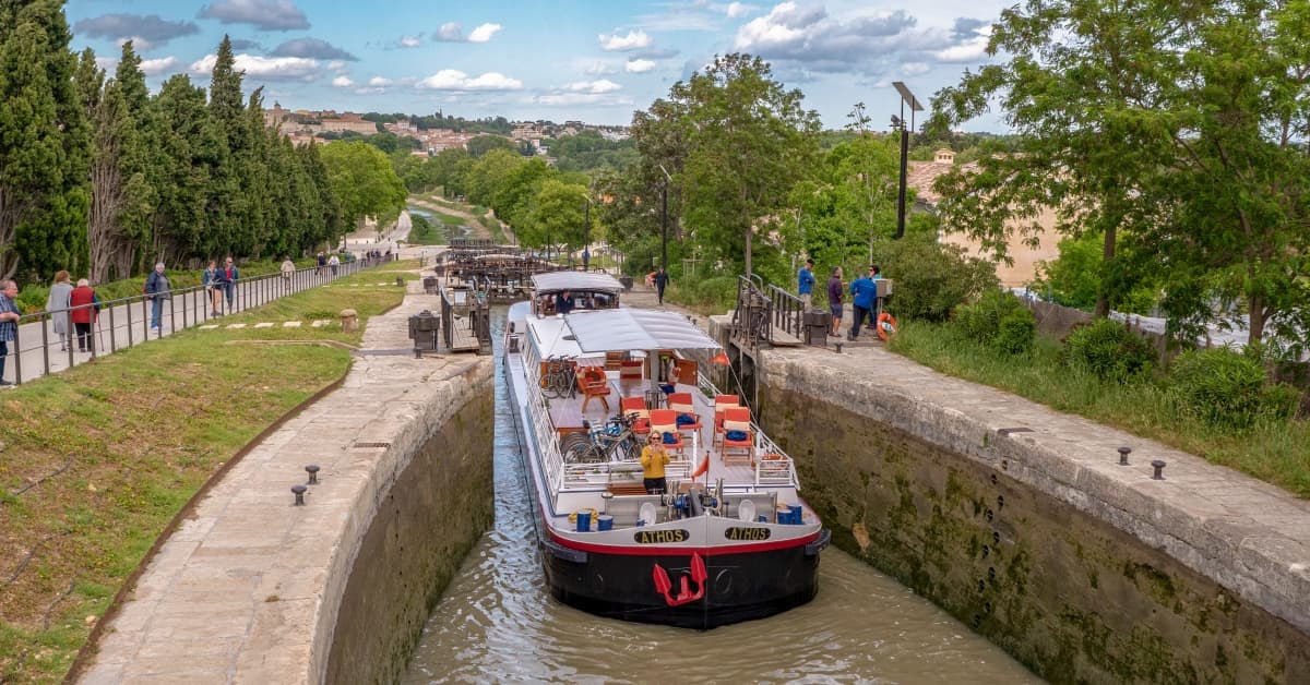 River Cruise Barge in European Canal Lock A red and white river cruise barge navigates through a stone lock system on a European waterway, with lush green banks, walking paths, and spectators observing the vessel's passage. The scenic landscape features historic architecture on hillsides in the background under a partly cloudy sky.