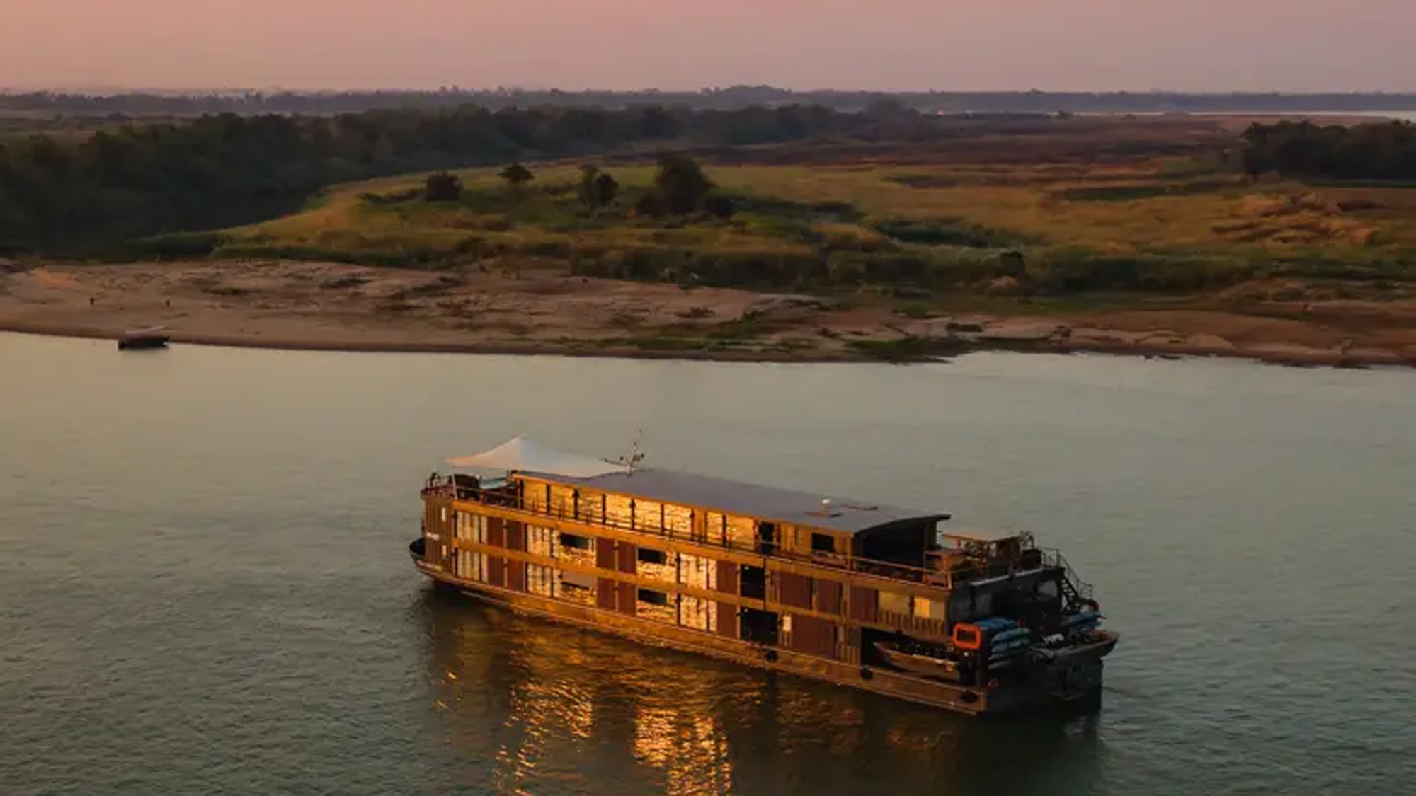 A traditional wooden river cruise ship navigates the Mekong River at sunset, with its distinctive multi-deck design illuminated by golden light. The vessel is positioned against a scenic backdrop of rural floodplain landscape with scattered trees and distant forests under a warm, hazy sky.
