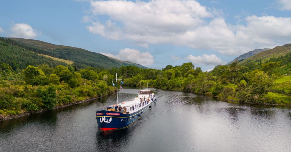 River Cruise Boat in Scottish Highlands A picturesque river cruise vessel navigates through calm waters surrounded by lush green mountains and forests in the Scottish Highlands. The scenic landscape features rolling hills, dense woodland, and dramatic mountain peaks under a partly cloudy sky, showcasing the natural beauty of Scotland's inland waterways.