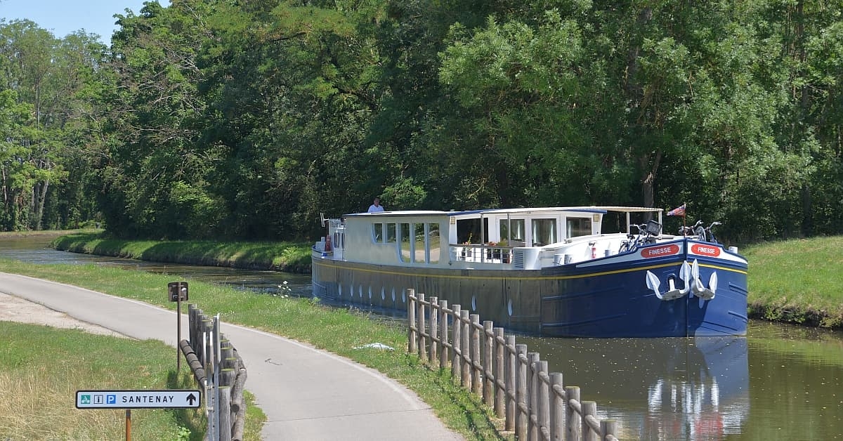 River Cruise Boat on European Waterway A modern river cruise vessel moored along a scenic European canal lined with lush green vegetation and towering trees. The white and blue boat features multiple passenger decks with numerous windows and appears to be docked at a designated port with wooden fencing and directional signage visible.
