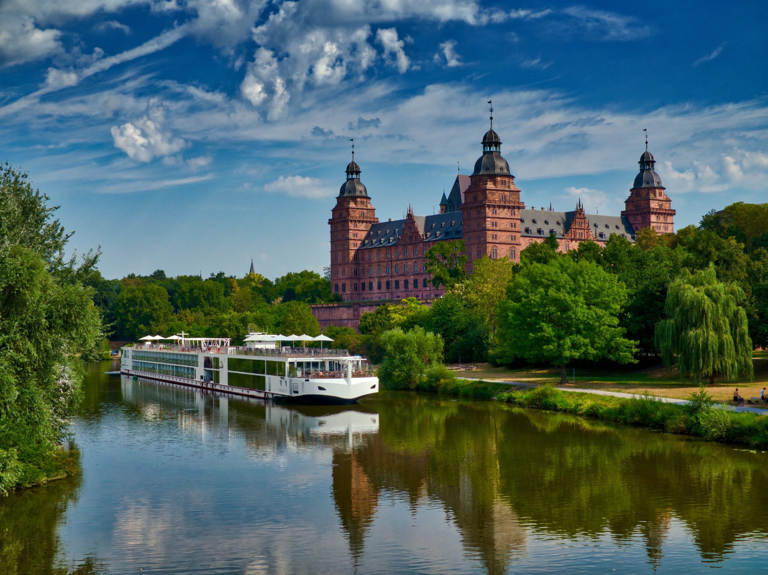 River Cruise Past Historic German Castle A modern river cruise ship navigates a tranquil river in front of an impressive Renaissance-style castle with multiple towers, surrounded by lush green trees and a dramatic blue sky with white clouds.