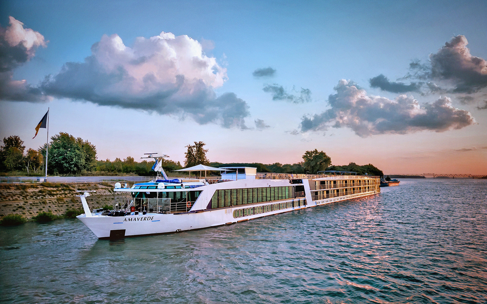 A white river cruise ship named Amaverde docked along a river at sunset, with dramatic cloudy skies and a tree-lined shore in the background. The vessel reflects on the calm water with soft golden and blue tones.