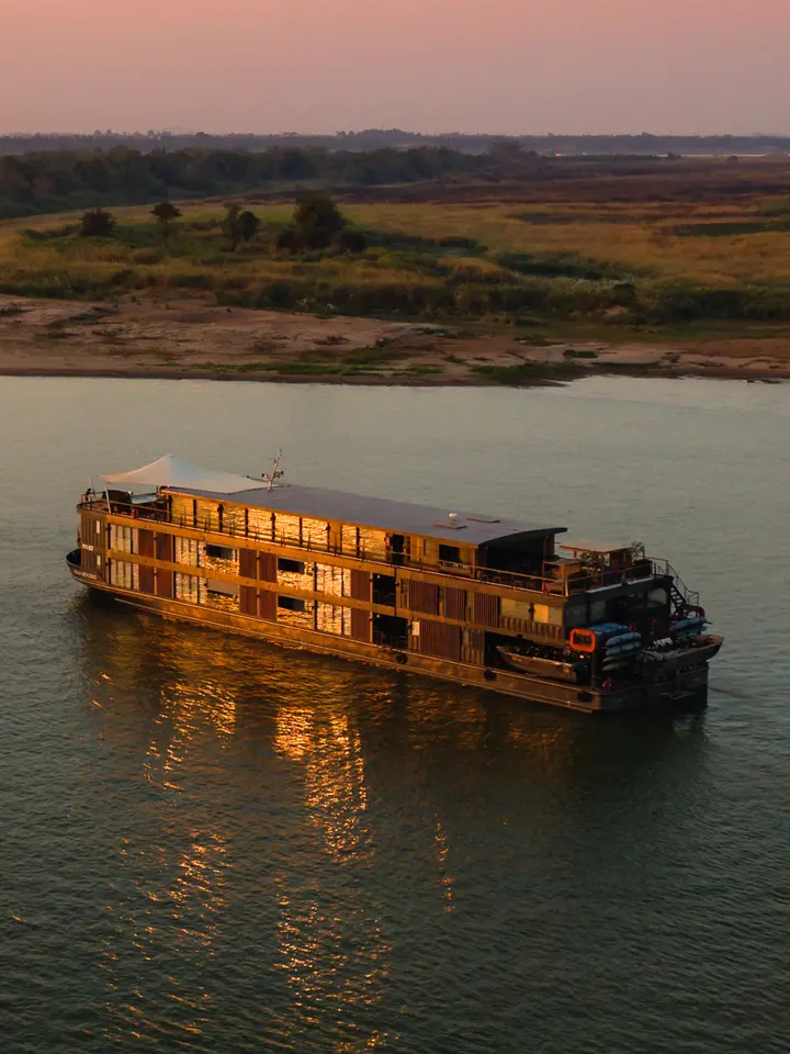 River Cruise Ship at Golden Hour A luxurious river cruise vessel with distinctive golden-lit wooden architecture floats peacefully on calm waters during sunset, with expansive floodplain landscapes and scattered trees visible along the distant riverbank under a warm, hazy sky.