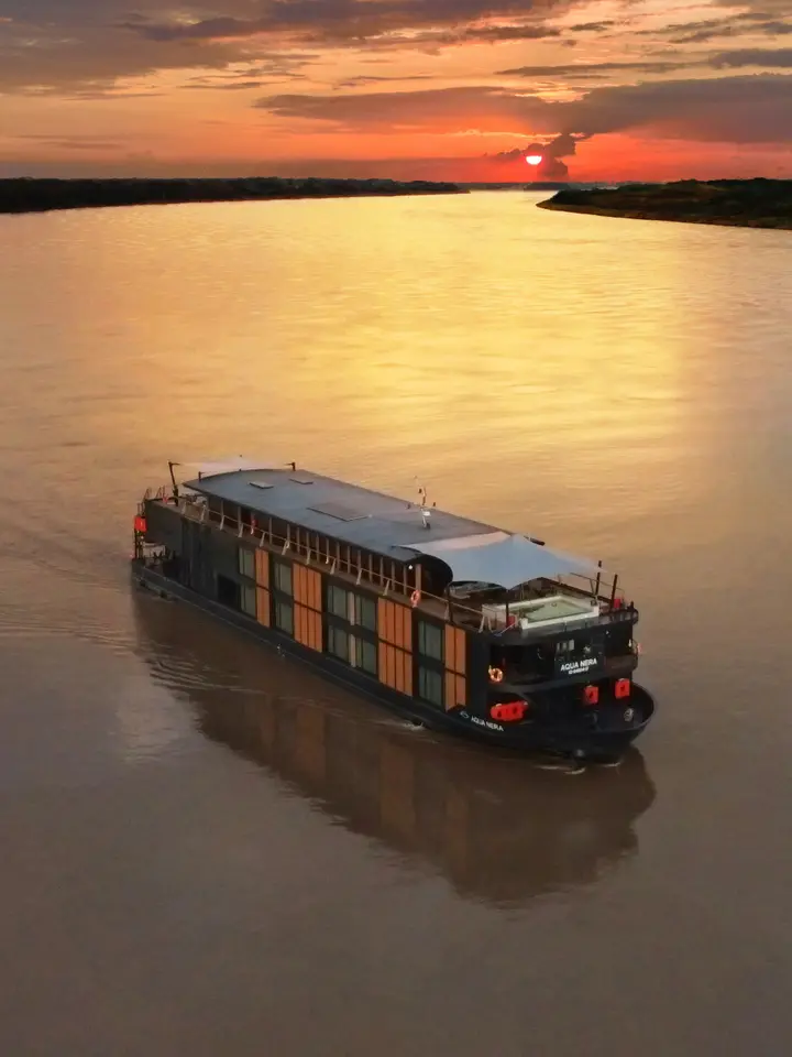 River Cruise Ship at Sunset A modern river cruise vessel with distinctive orange and dark hull coloring navigates a calm, wide river during a spectacular golden sunset. The ship's reflection mirrors in the still water as the sun dips below the horizon, creating a serene travel scene typical of river cruising in Southeast Asia or similar riverine destinations.