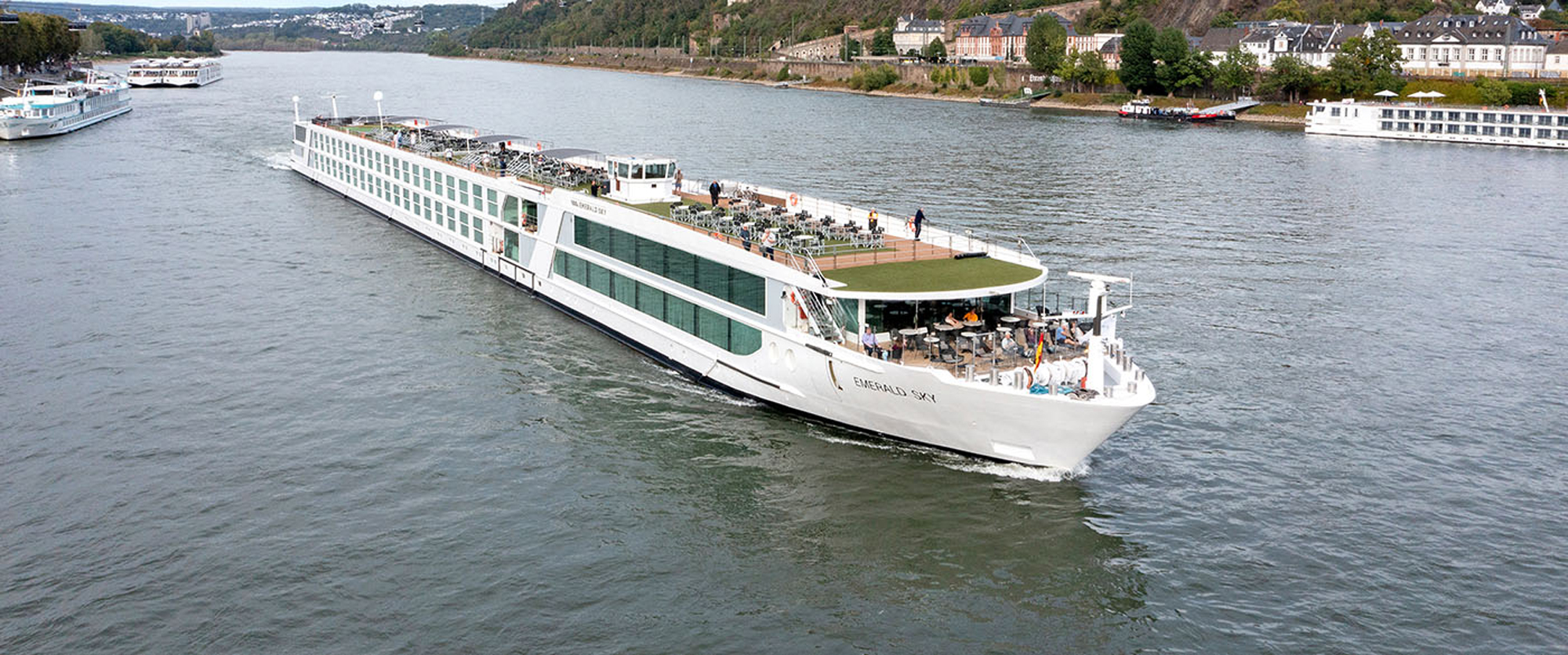 River Cruise Ship on European Waterway A modern white river cruise ship sailing along a scenic river with historic buildings and landscape in the background. The vessel features multiple decks with passengers enjoying the journey.