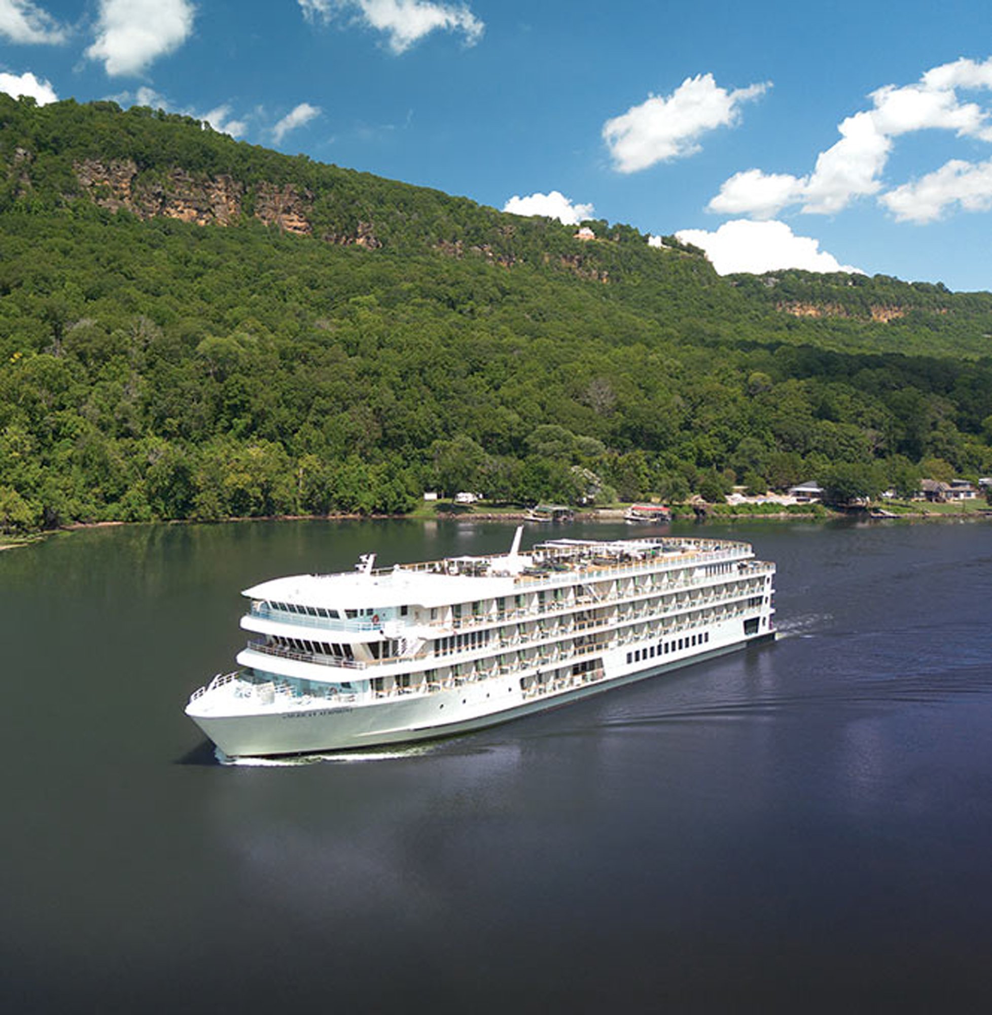A large white river cruise ship navigates a calm river flanked by lush, densely forested hillsides with exposed rocky outcrops. The vessel features multiple passenger decks with balconies and is positioned against a picturesque landscape with charming riverside buildings visible along the shoreline.