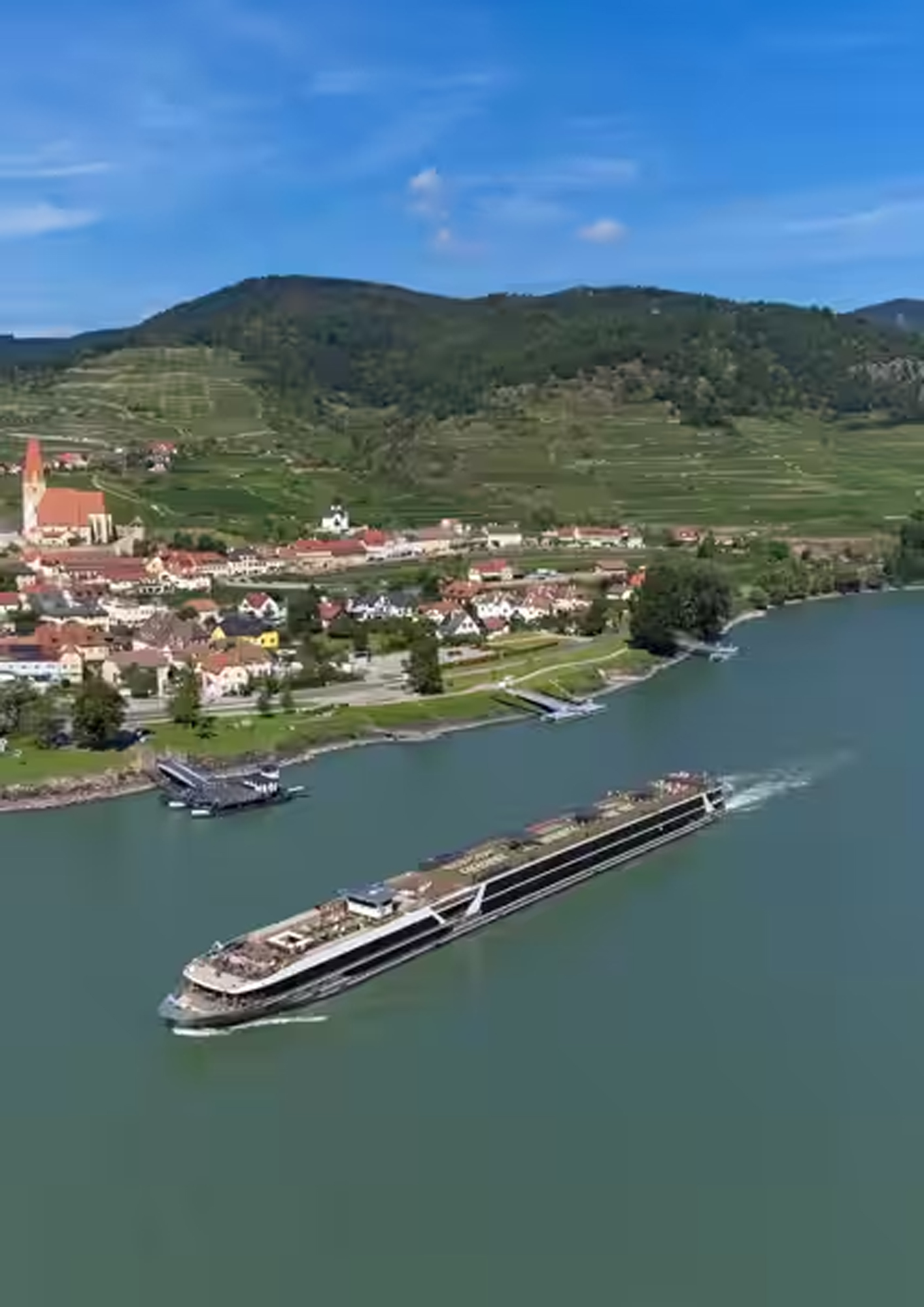 River Cruise Through European Vineyard Landscape A modern river cruise ship sailing along a picturesque waterway with a charming European village nestled on the shoreline, surrounded by terraced vineyards and forested hills.