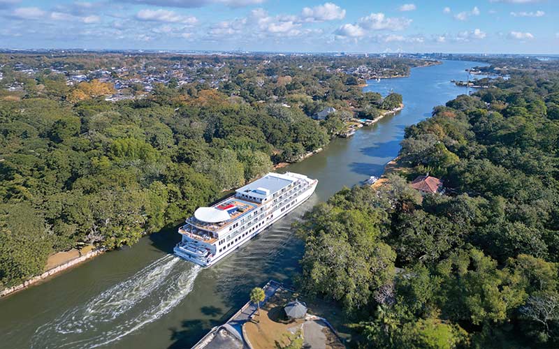 River Cruise Through Lush Landscape A large white river cruise ship navigating through a verdant waterway surrounded by dense trees and coastal urban development. The vessel moves smoothly through the calm waters with forested banks on both sides.