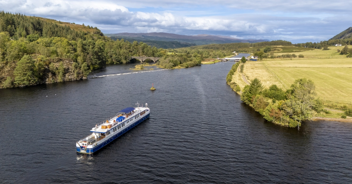 River Cruise Vessel on Scottish Waterway A scenic river cruise ship navigates through a picturesque Scottish valley, surrounded by lush forests, rolling hills, and pastoral farmland. The vessel glides peacefully along the water with a historic stone bridge visible in the distance and mountains framing the horizon.