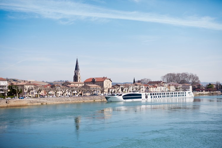 A white river cruise ship docked alongside a picturesque European town with a prominent church spire, situated on a calm blue river with historic buildings in the background.