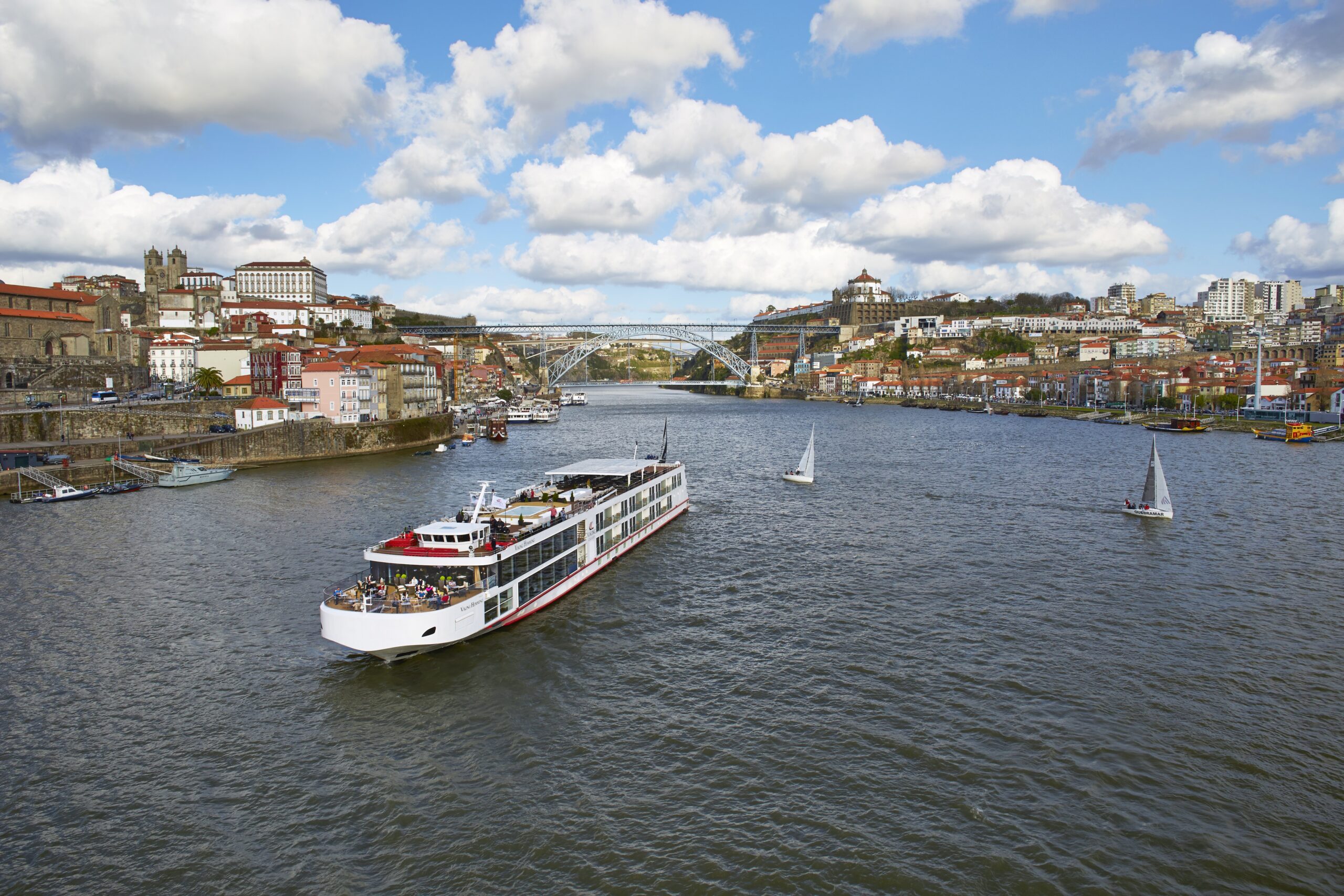 River Cruise in Porto, Portugal A modern river cruise ship navigates the Douro River in Porto, with the iconic Dom Luís I Bridge and historic city architecture in the background. Two sailboats are also visible on the river.