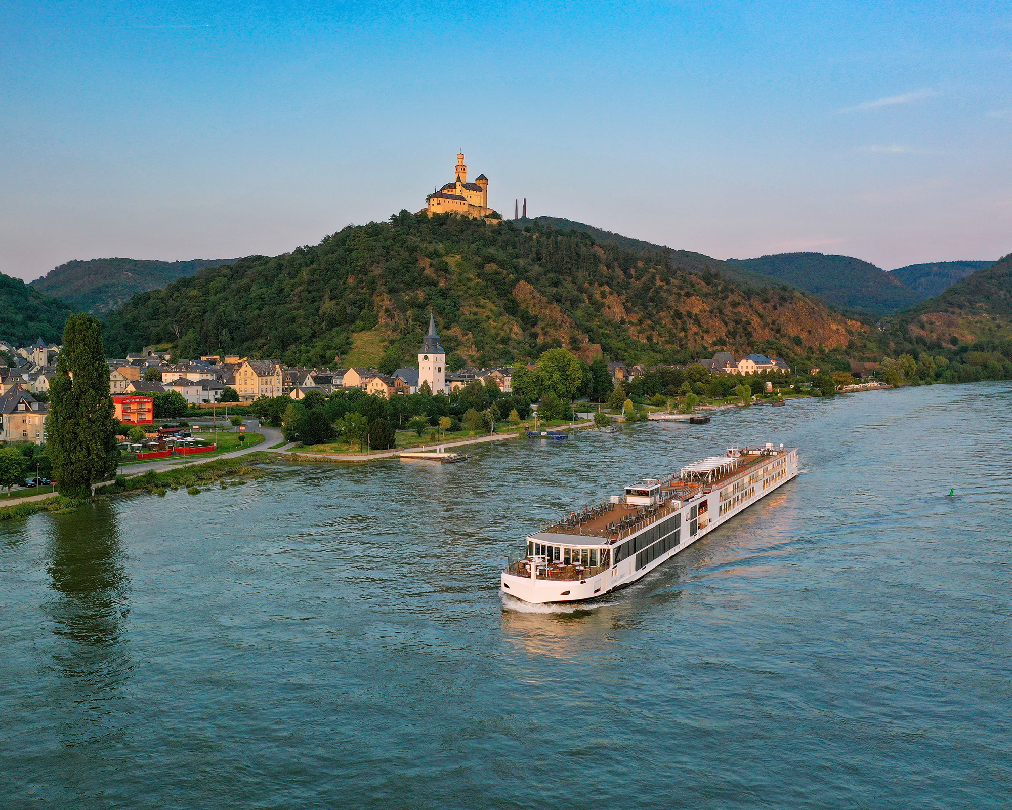 River Cruise on the Moselle in Germany A modern river cruise ship sailing along the Moselle River, with a picturesque medieval castle perched on a forested hill and a charming European town in the foreground.