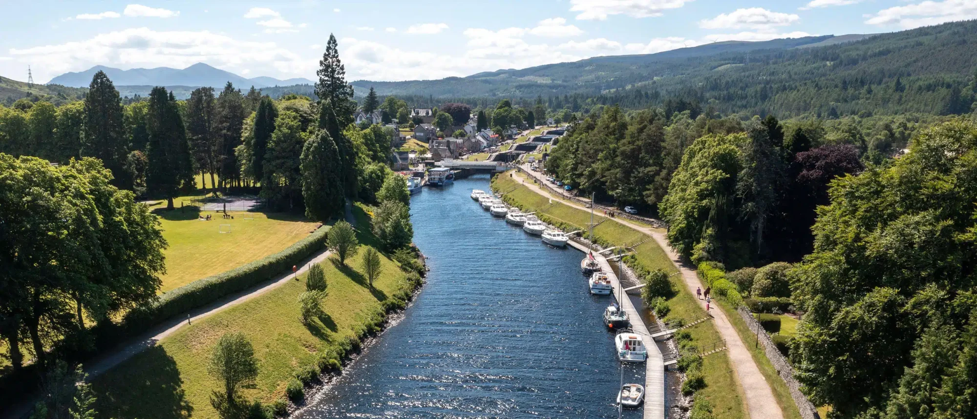 River Town with Moored Boats and Mountain Views An idyllic riverside town nestled in a forested valley with numerous pleasure boats and small vessels moored along manicured banks, surrounded by dense coniferous forests and distant mountains under a partly cloudy sky. The scene captures a charming European river destination with elegant waterfront promenades and traditional architecture.