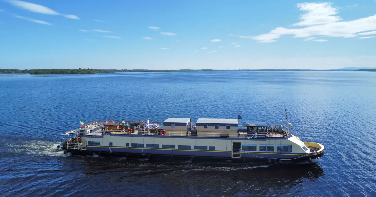 Riverboat Cruise Ship on Scenic Lake A multi-deck riverboat or lake cruise vessel anchored in calm blue waters with forested shoreline visible in the distance under clear skies. The ship features a cream and blue color scheme with multiple passenger decks, outdoor recreational areas on the upper deck, and appears to be a regional or inland waterway cruise vessel.