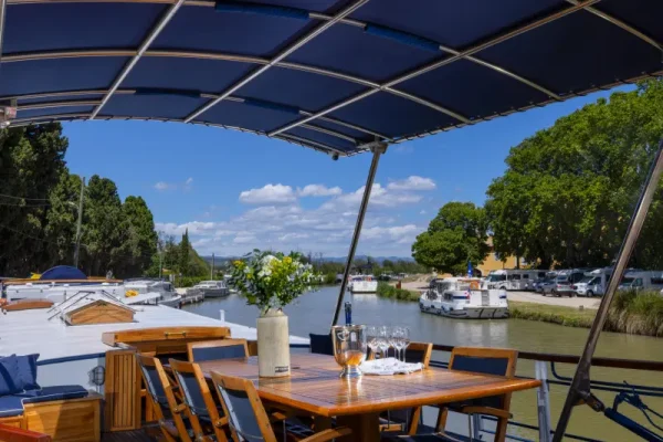 Riverside Dining Deck with Solar Canopy An elegant outdoor dining area situated on a river cruise vessel, featuring a wooden table set for dining under a blue solar panel canopy, with scenic riverside views of moored boats, lush green vegetation, and distant mountains visible beyond the waterway.
