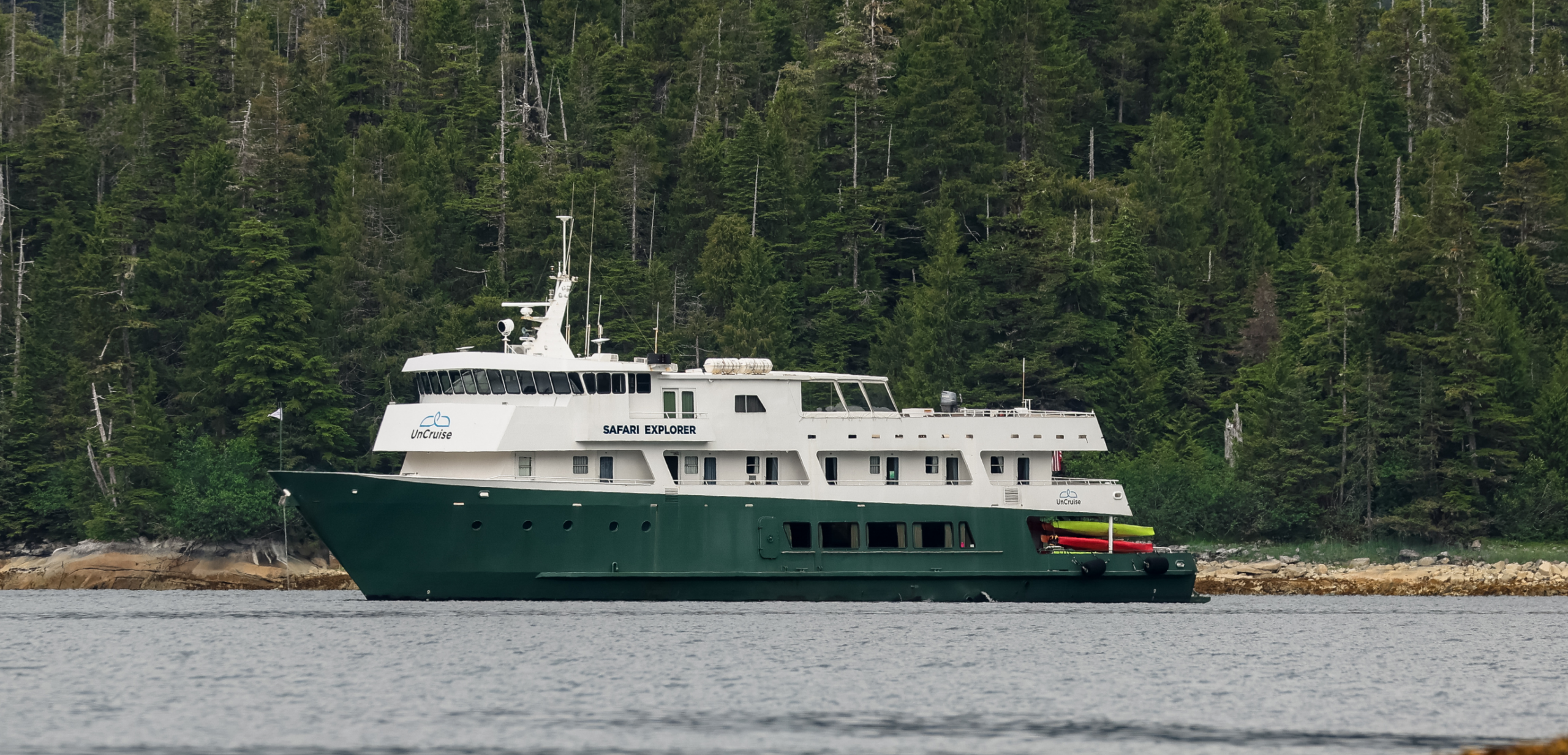 Safari Explorer Vessel in Coastal Forest A green-hulled expedition cruise ship named Safari Explorer anchored near a rocky shoreline with dense evergreen forest in the background. The vessel appears equipped for wilderness exploration with kayaks visible on its deck.