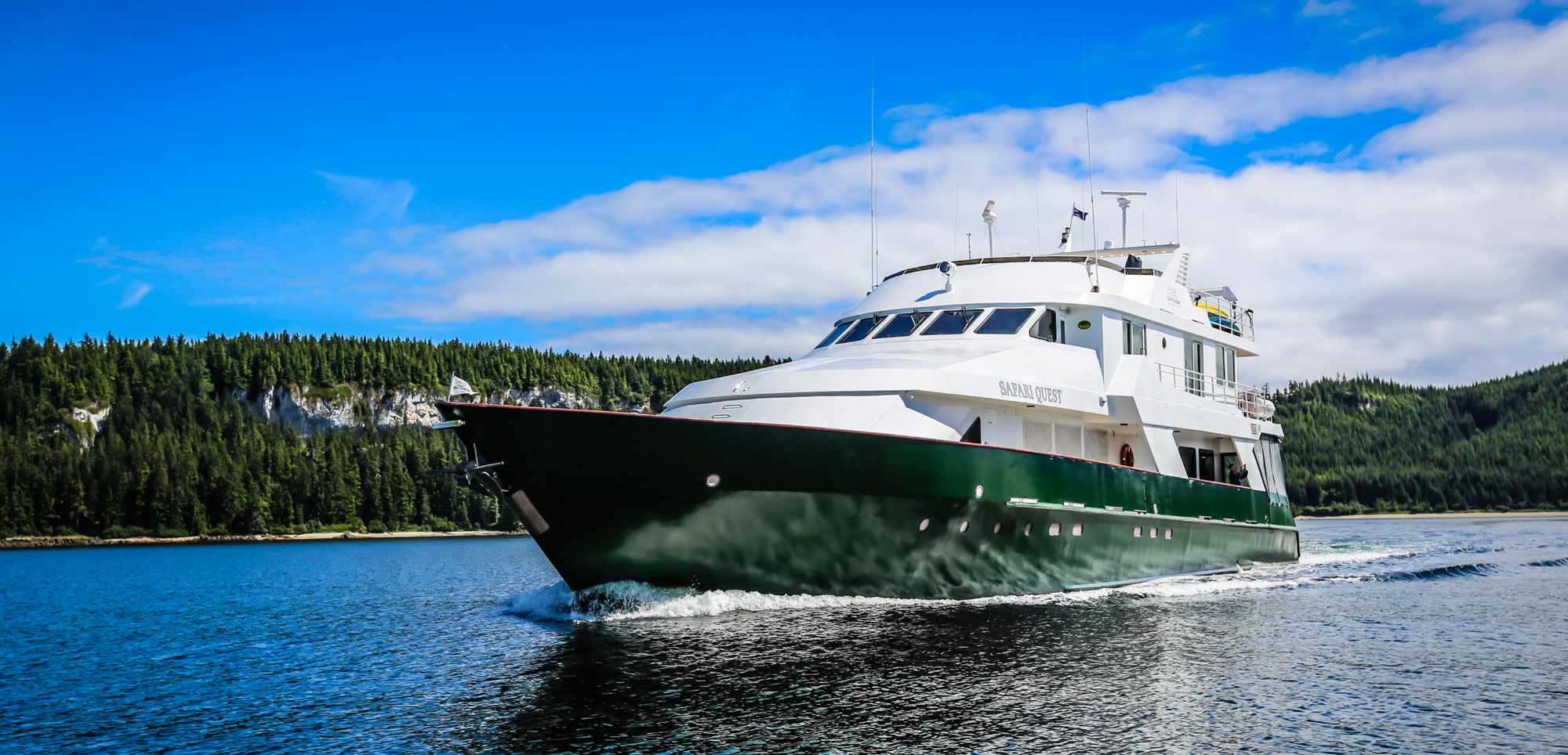 Safari Quest Expedition Vessel in Forested Waterway A white and green expedition cruise ship named Safari Quest navigating through a scenic forested waterway with dense evergreen forests and rocky shorelines in the background.