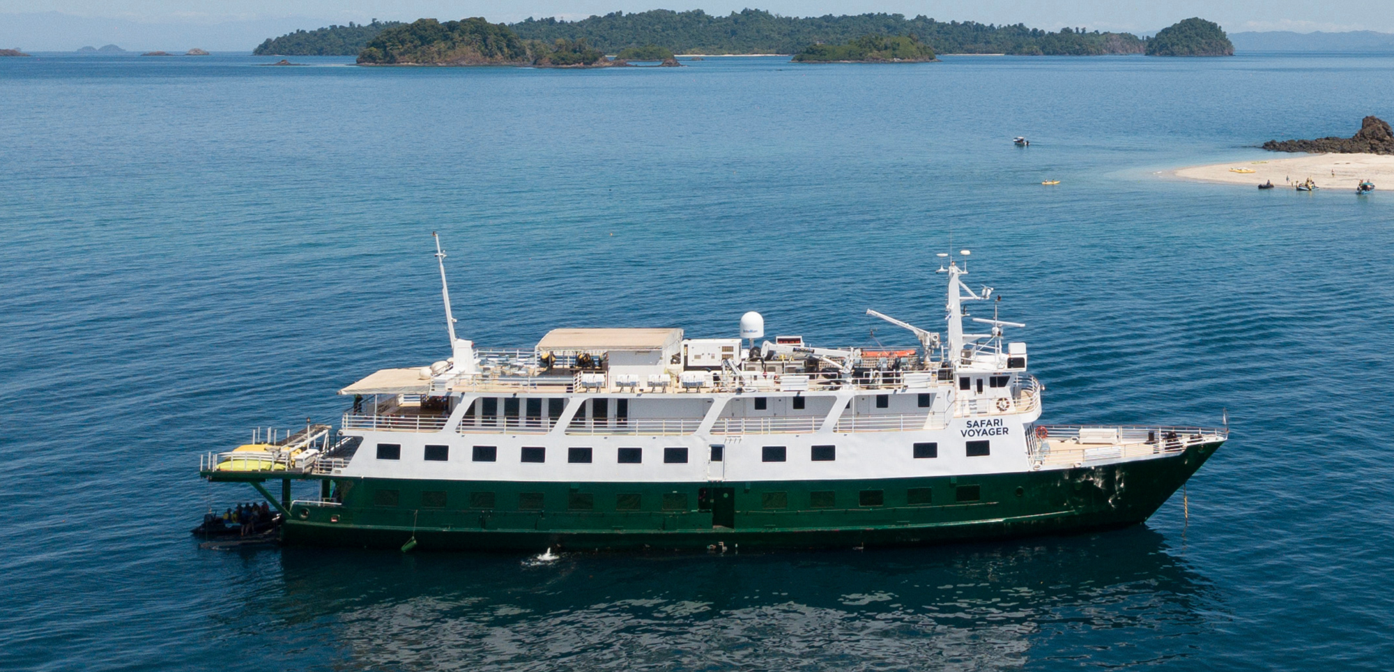 Safari Voyager Expedition Cruise in Tropical Waters A green and white expedition cruise ship anchored in calm blue waters with lush tropical islands in the background. The Safari Voyager appears to be a small adventure cruise vessel positioned near a sandy beach and scenic archipelago.