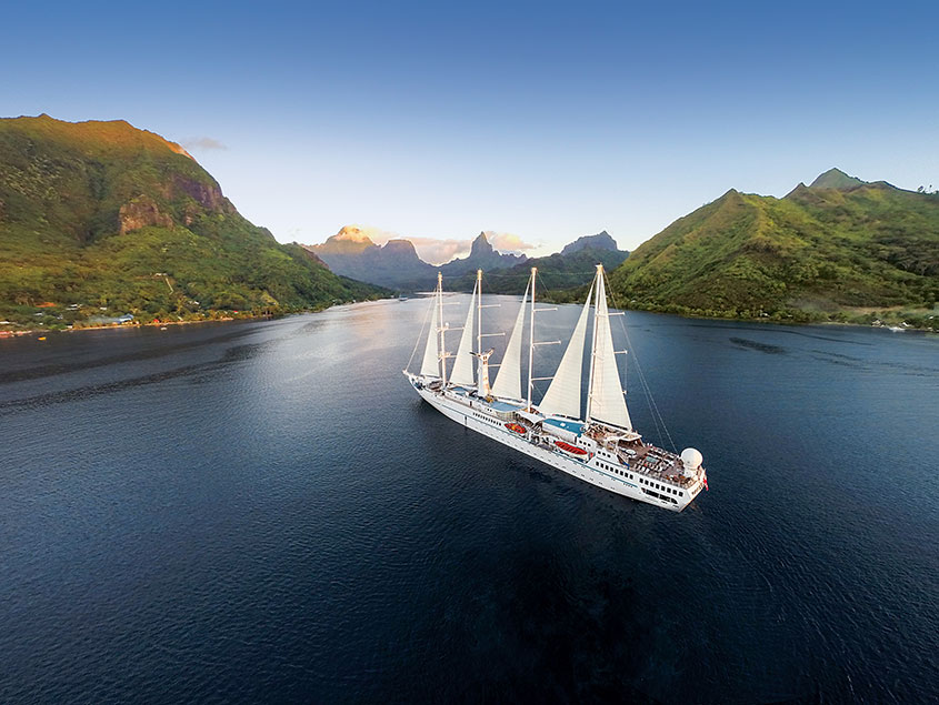 A white multi-masted sailing vessel navigating through a scenic waterway surrounded by lush green mountainous terrain with dramatic peaks and coastline.