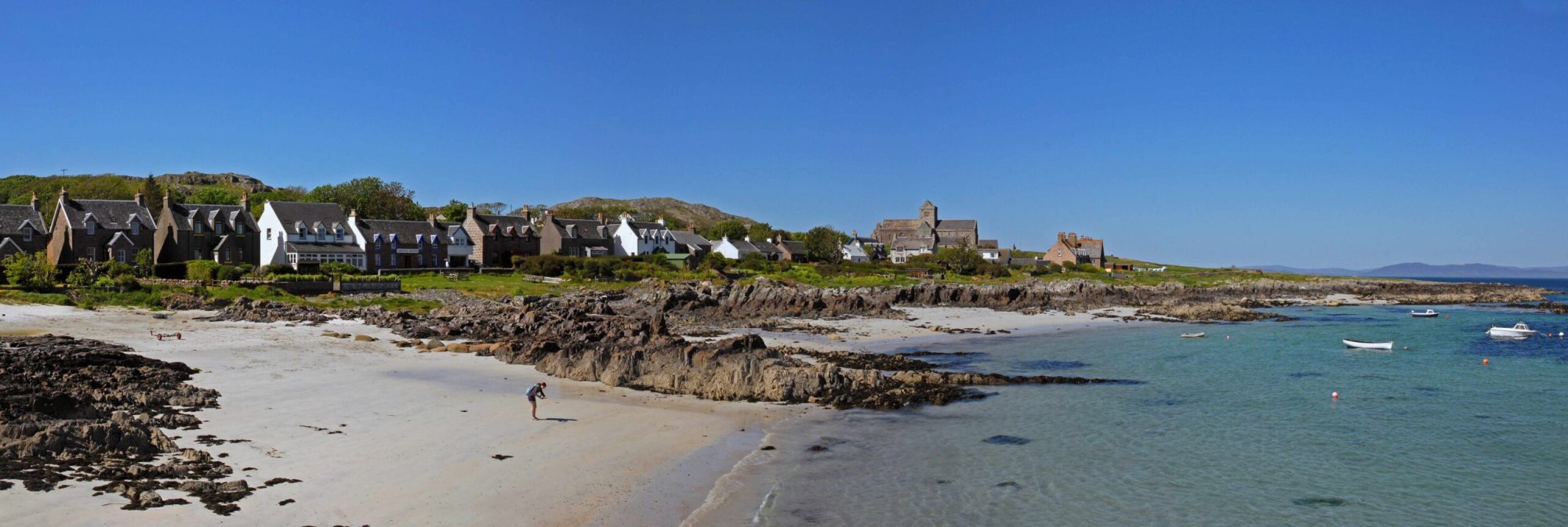 Scottish Coastal Village with Beach and Boats A picturesque Scottish coastal settlement featuring traditional stone architecture with distinctive white-trimmed buildings nestled against rolling hillsides, overlooking a pristine sandy beach dotted with moored boats and rocky outcrops in turquoise waters. The scene captures the rugged beauty of Scotland's northern coast with its characteristic blend of natural landscape and charming seaside village character.