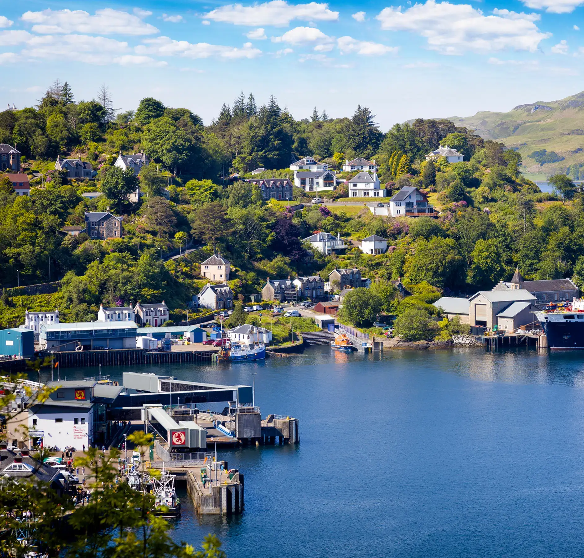 Scottish Highland Village Harbor View A picturesque Scottish waterfront village nestled in a lush valley with traditional stone buildings, modern homes scattered across tree-covered hillsides, and a working harbor with fishing boats and maritime facilities. The serene blue waters reflect the verdant landscape under a bright summer sky, capturing the charm of a remote Highland coastal community.