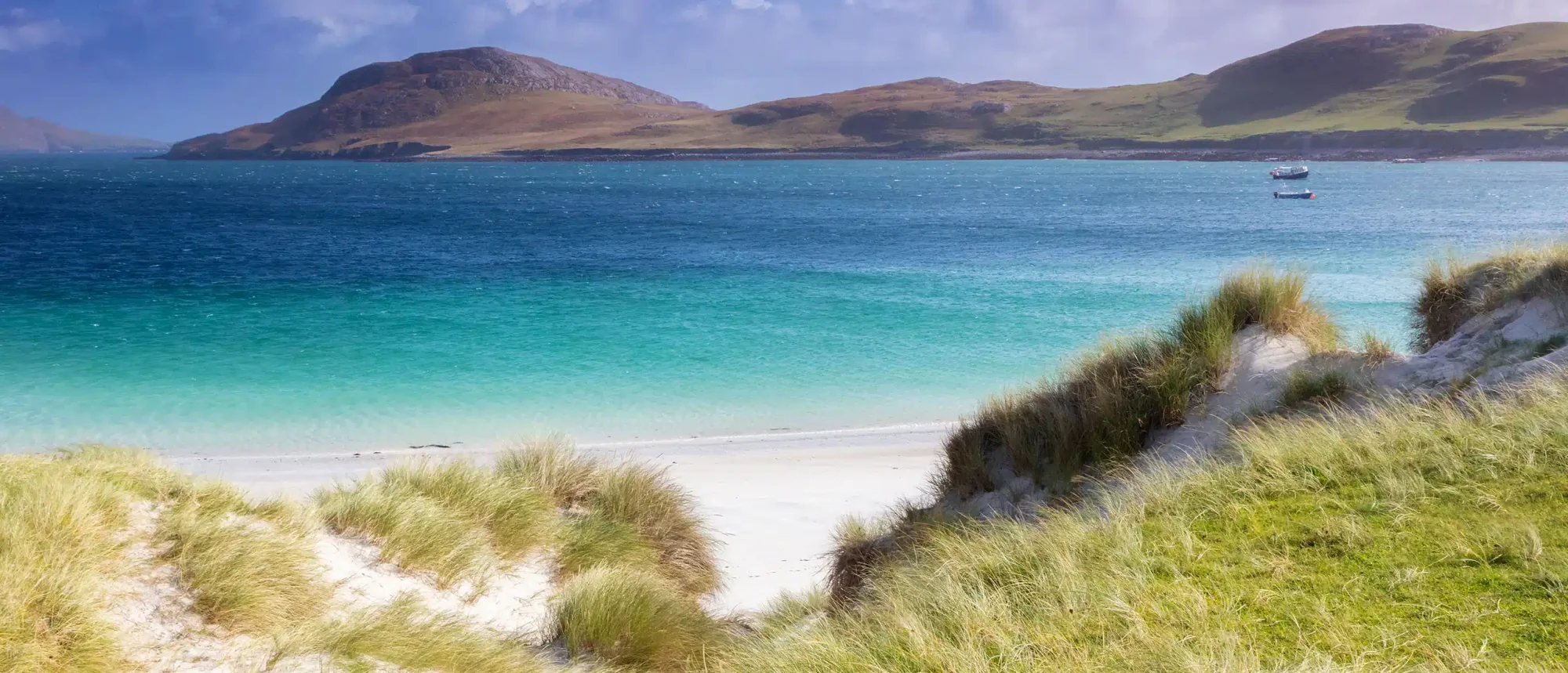 Scottish Island Beach with Turquoise Waters A pristine white sand beach with coastal dune grasses overlooks crystalline turquoise waters and rolling green hills across the bay, featuring traditional fishing boats anchored in the calm waters. This remote Scottish island destination showcases dramatic mountainous terrain and exemplifies the rugged natural beauty characteristic of the Hebrides.
