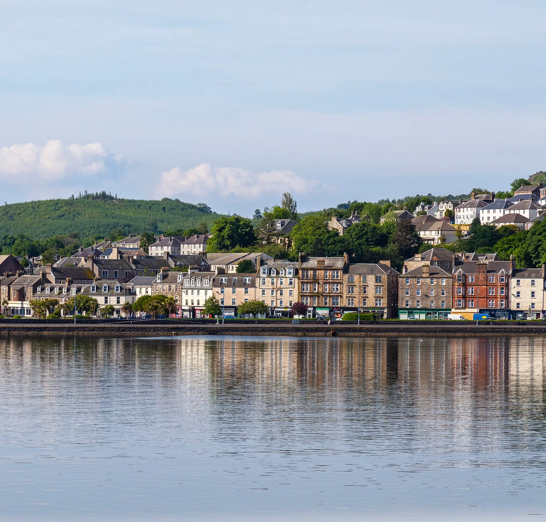 Scottish Riverside Town Waterfront View A picturesque Scottish riverside town featuring a row of traditional Georgian and Victorian stone buildings reflected in calm waters, with forested hills rising in the background. The charming waterfront townscape with its varied architecture and natural setting exemplifies classic Scottish Highlands destinations popular with cruise itineraries.