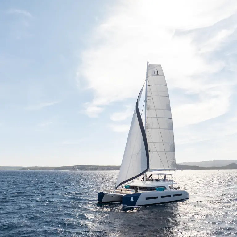 Sleek White Catamaran Sailing Blue Waters A modern white catamaran with fully raised sails navigating through bright blue ocean waters with distant coastline in the background. The vessel appears to be a luxury sailing yacht with clean, contemporary design.
