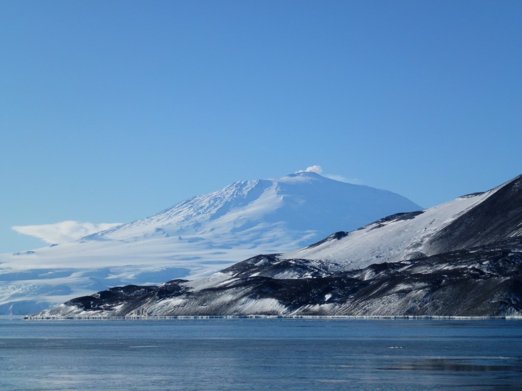 Snow-Capped Volcano Above Frozen Lake A majestic snow-covered volcanic peak rises dramatically above a frozen lake with dark volcanic rock formations along the shoreline, captured under clear blue skies in what appears to be a polar or sub-arctic region.