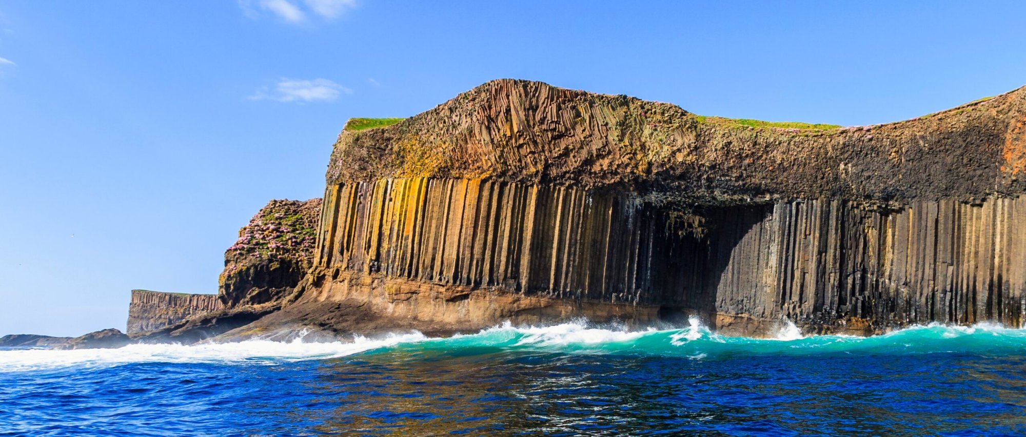 Staffa Basalt Columns and Coastal Cliffs A dramatic coastal landscape featuring distinctive hexagonal basalt columns rising from turquoise waters, with waves crashing against the volcanic rock formations. The scene showcases the UNESCO-recognized geological formations of Staffa in Scotland, a popular destination for cruise ship excursions.