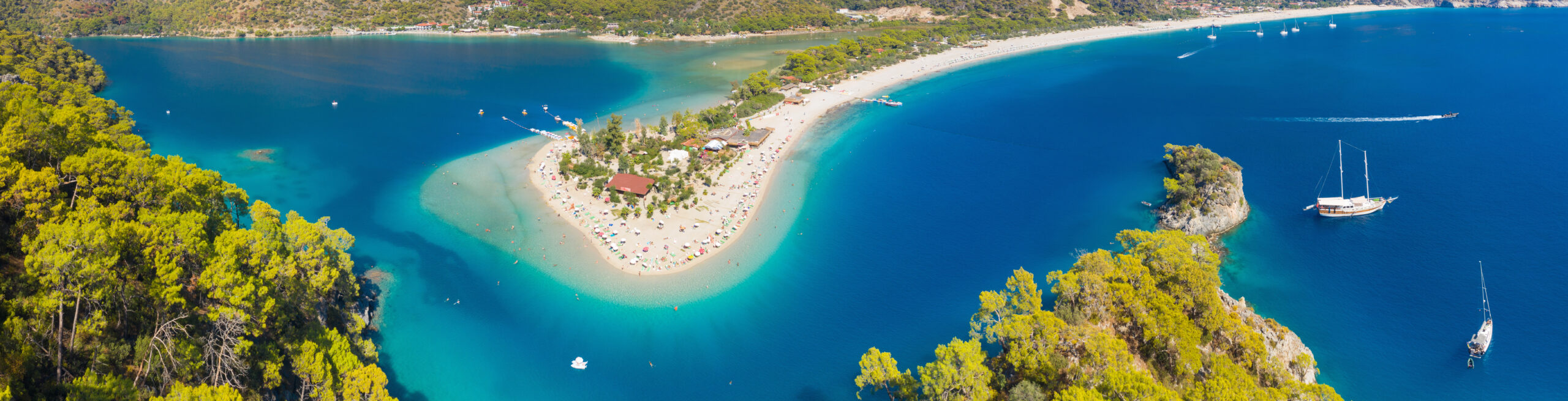 An aerial view of a unique sandy peninsula with white beaches extending into turquoise blue waters, surrounded by green forested coastline and featuring several boats and sailboats.