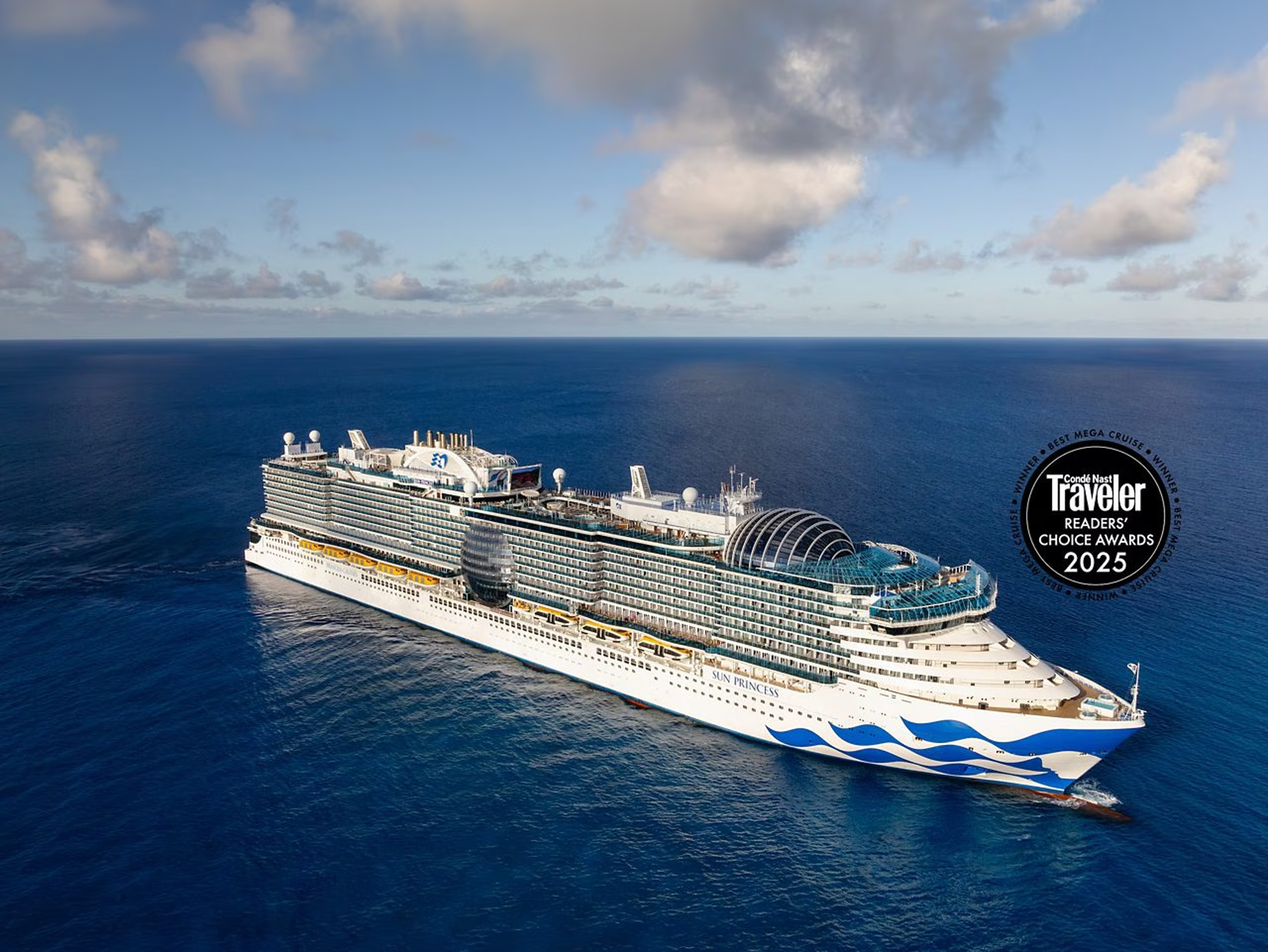 A large modern cruise ship sailing on deep blue ocean waters under a cloudy sky, featuring a sleek white and blue design with multiple decks and a distinctive glass dome structure.