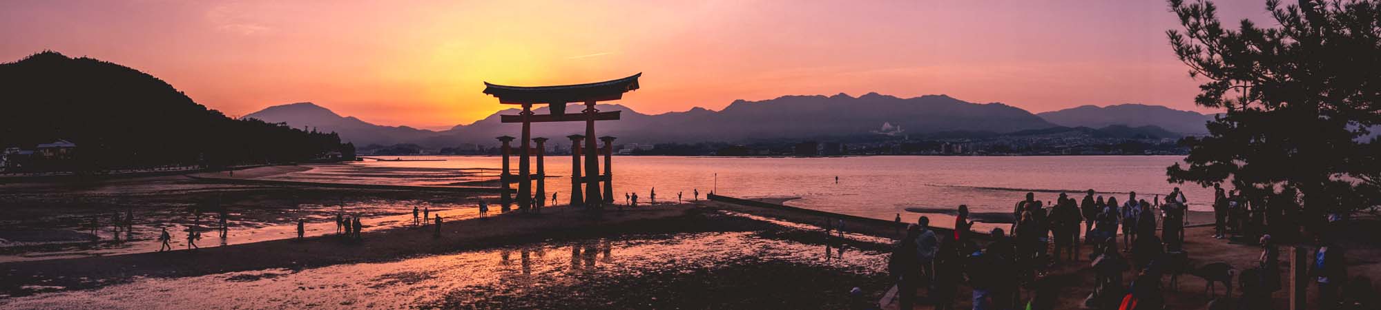 Sunset at Miyajima’s Famous Torii Gate A stunning sunset view of the iconic Itsukushima Shrine's floating torii gate at low tide, with silhouetted visitors and mountains in the background.