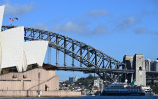 A panoramic view of Sydney Harbour featuring the Sydney Opera House and the Sydney Harbour Bridge, with a cruise ship in the foreground and the city skyline in the background.