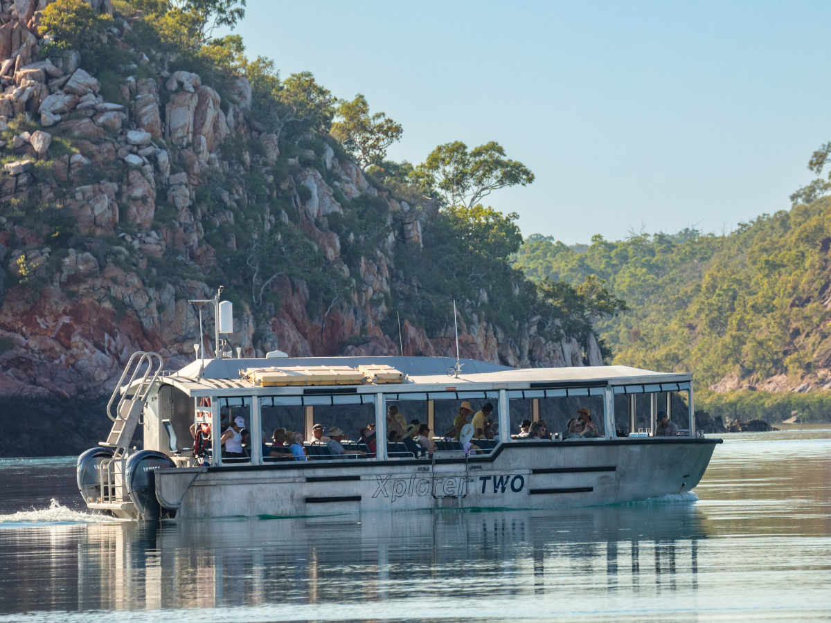 A tour boat named 'Xplorer Two' navigating through a scenic waterway with rocky cliffs and lush vegetation in the background. The boat is filled with passengers enjoying a scenic cruise.
