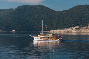 Traditional Gulet Sailing in Turkish Waters A classic wooden gulet sailing vessel with traditional Turkish design anchors in calm, crystal-clear blue waters surrounded by forested mountains. The boat features distinctive twin masts and golden-toned wooden construction, reflecting in the mirror-like sea beneath clear skies.