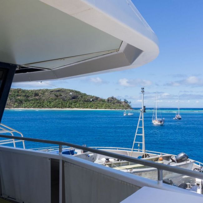 Tropical Island Anchorage with Sailboats A scenic view from a cruise ship's deck showing multiple sailboats anchored in crystal-clear turquoise waters off a pristine tropical island with white sandy beaches and lush green vegetation. The image captures a perfect tropical paradise destination with clear blue skies and ideal sailing conditions.