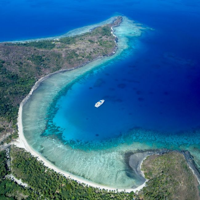 Tropical Island Paradise Aerial View An aerial view of a pristine tropical island featuring a crescent-shaped white sand beach, turquoise shallow waters, and dense green vegetation, with a single white yacht anchored in the crystal-clear lagoon. The vibrant gradient of blues and greens showcases the shallow reef waters contrasting with the deeper ocean.