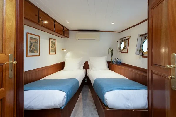 Twin Bed Cabin Aboard Cruise Ship A compact twin-bed cabin aboard a cruise ship featuring two single beds with blue covers, wooden cabinetry, white walls, and nautical-style portholes. The space exemplifies efficient cruise ship accommodation design with warm wood tones and practical furnishings typical of mid-range cruise vessel interiors.