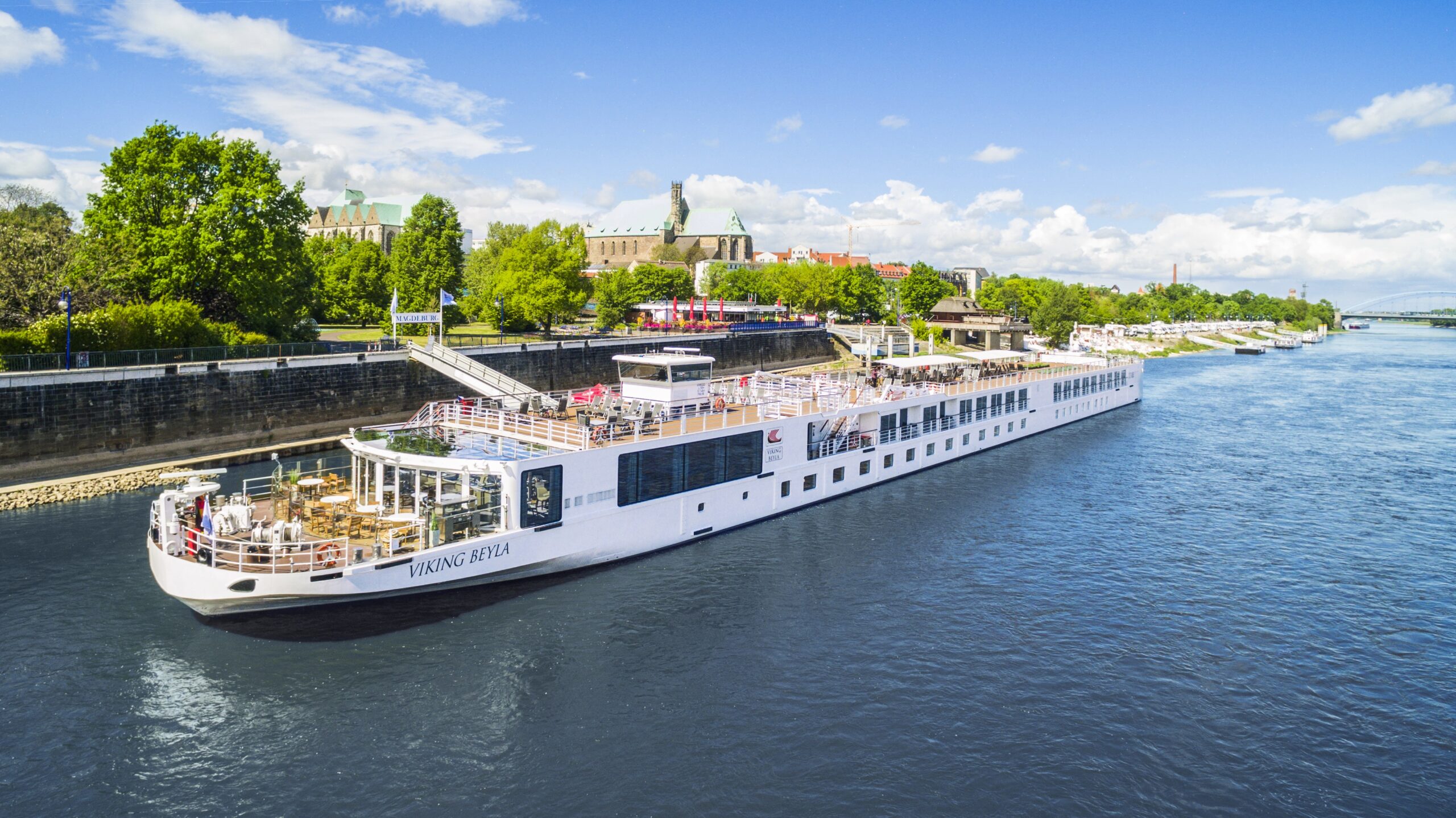 Viking River Cruise on European Waterway A white Viking river cruise ship docked near a historic riverside city with green trees and architectural landmarks. The vessel is moored in a lock with a picturesque urban landscape in the background.