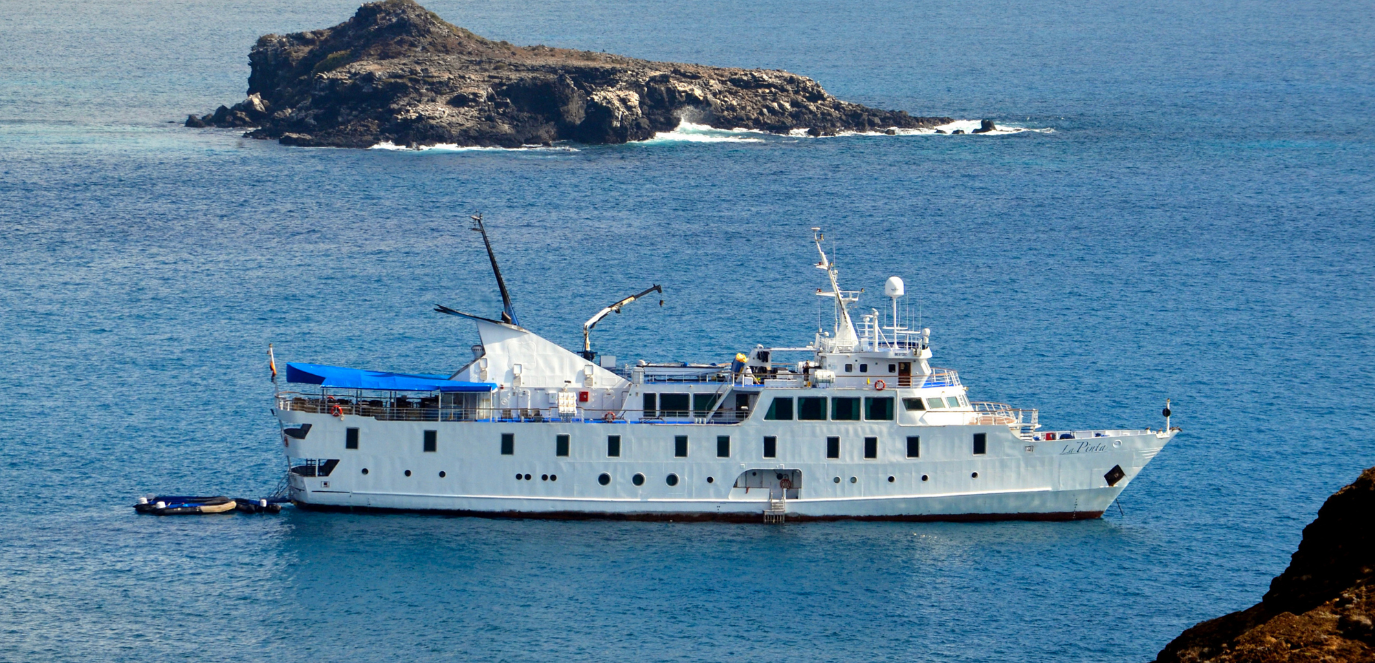 White Cruise Ship Near Rocky Island A white expedition-style cruise vessel anchored near a rocky, volcanic island with blue ocean waters surrounding it. The ship appears to be a small, specialized cruise boat with multiple decks and navigation equipment.